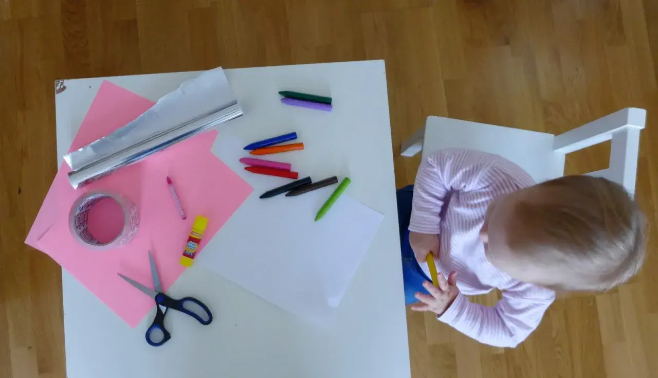 Dziecko przygotowuje materiały do zrobienia lustra z folii aluminiowej: kredki, nożyczki, taśmę, papier i folię.