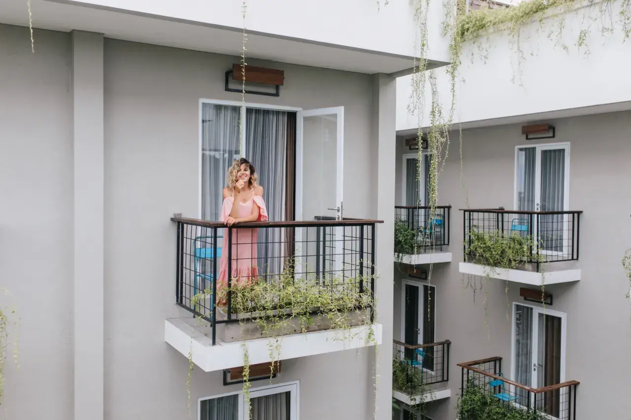 Kobieta w różowej sukience stoi na balkonie, opierając się o balustradę. Wokół rosną zielone rośliny, tworząc naturalną zasłonę na balkon.