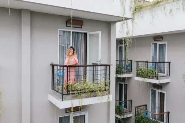 Kobieta w różowej sukience stoi na balkonie, opierając się o balustradę. Wokół rosną zielone rośliny, tworząc naturalną zasłonę na balkon.